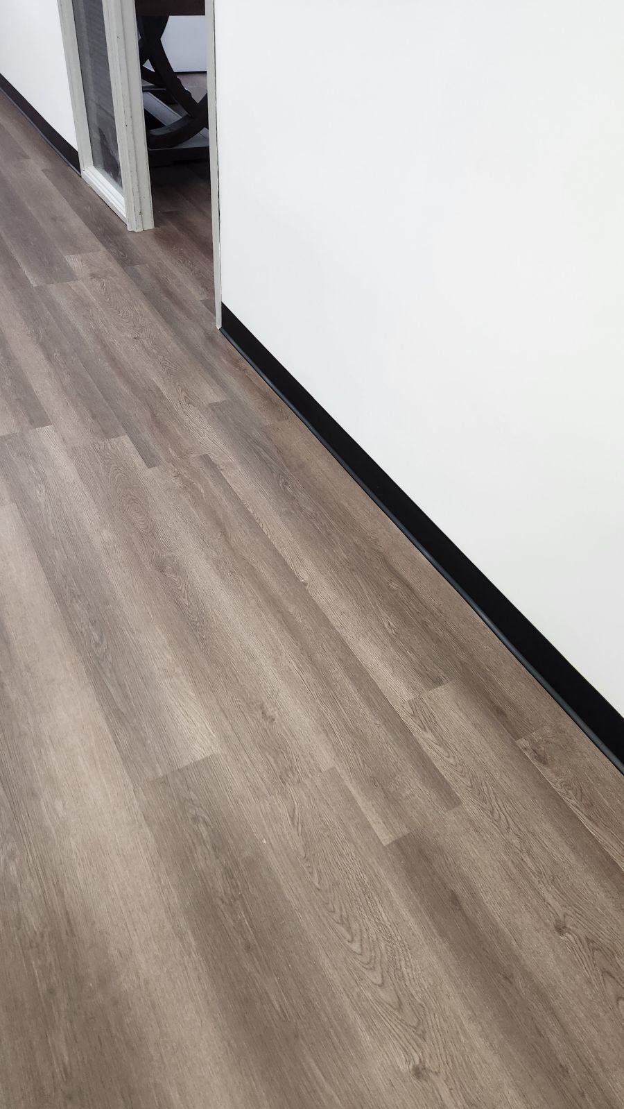A hallway with light brown wood-look flooring, a white wall, and a black baseboard, leading to a door.