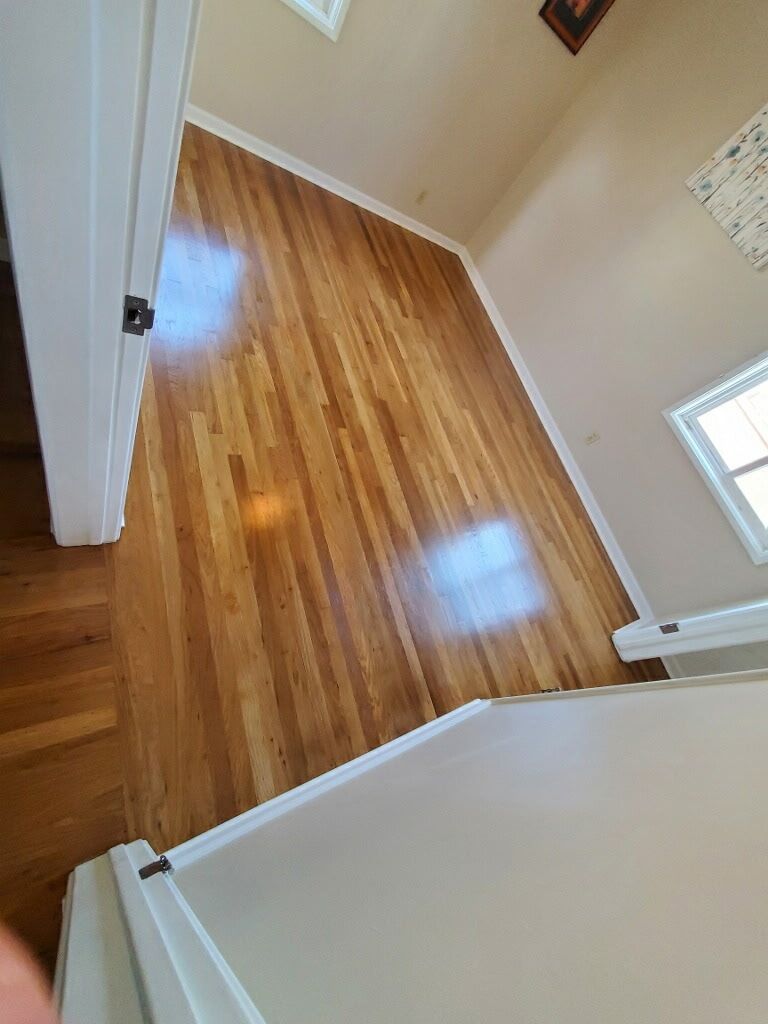 Empty room with polished light-brown hardwood floors and beige walls, viewed through an open doorway.