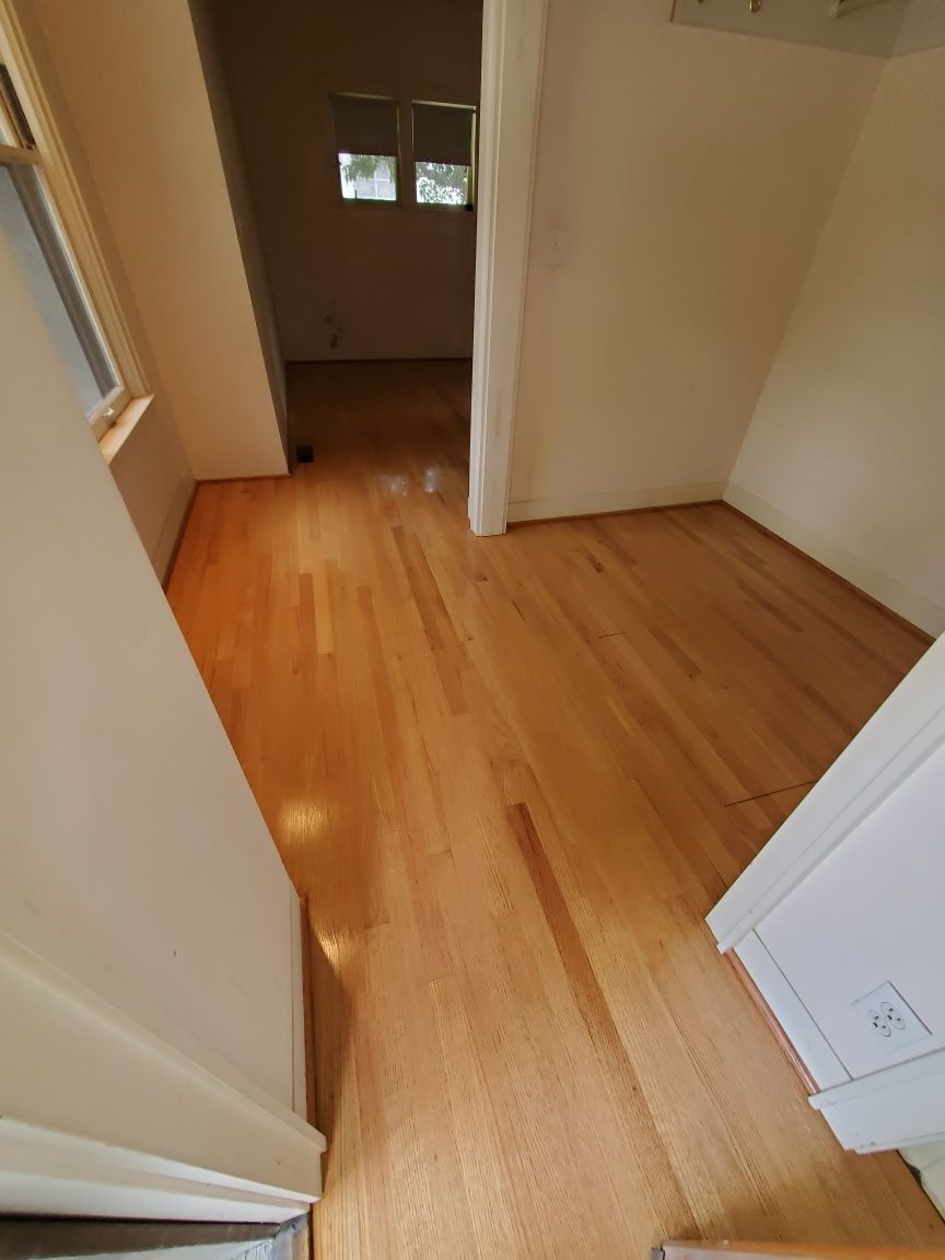 A view of a brightly lit room with light-colored hardwood floors, white walls, and a doorway leading to another room.