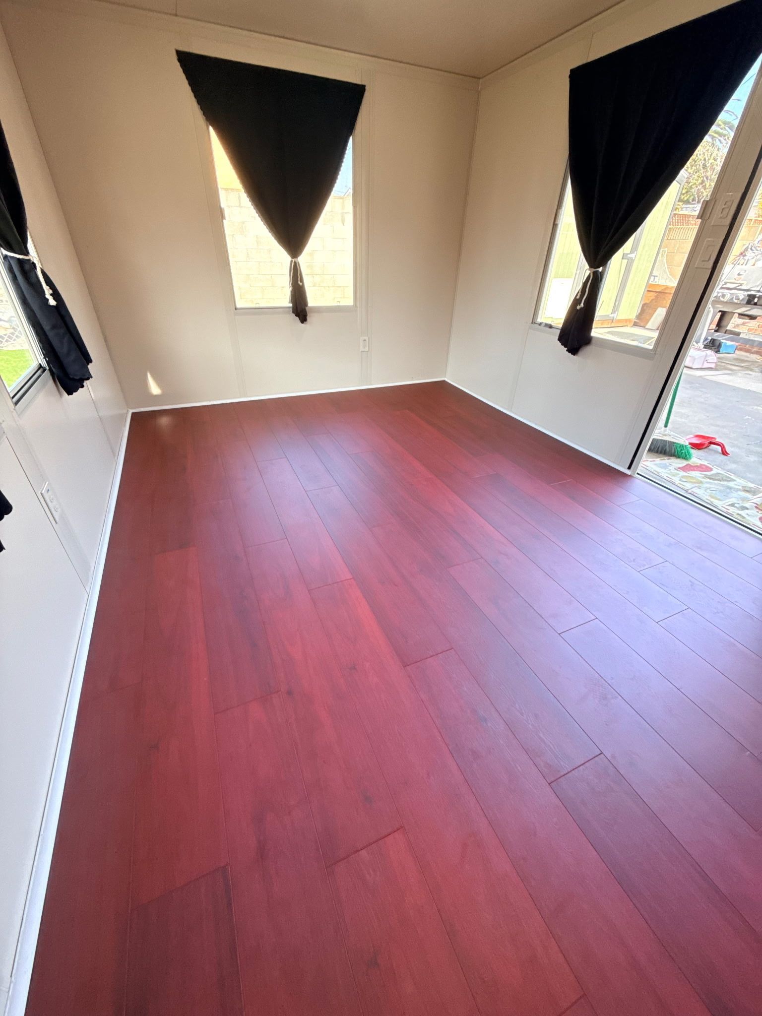 An interior view of an empty room with deep reddish-brown wood flooring, off-white walls, and black triangular curtains.