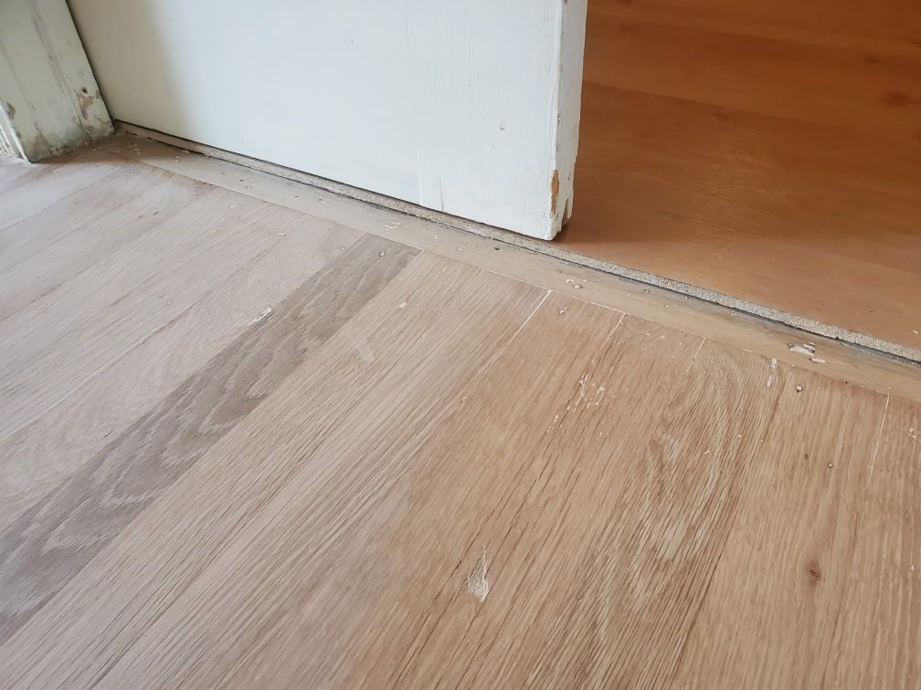 A transition threshold between two different types of light-colored hardwood floors at a doorway.