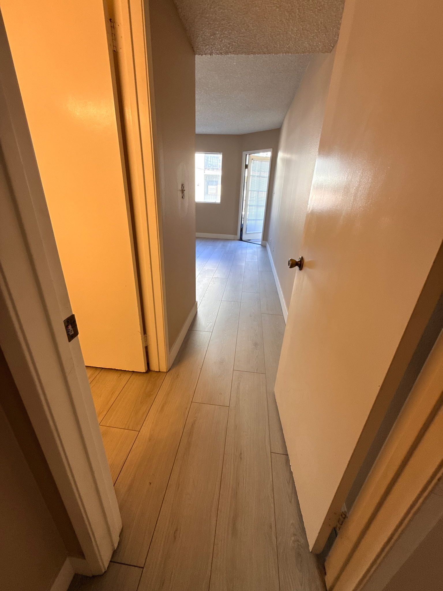 A narrow hallway with light-toned wood-plank flooring leading to an open doorway with a view of a window.