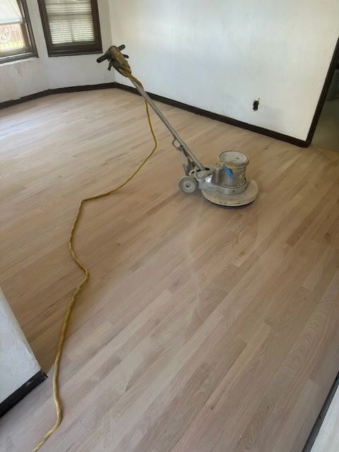 A floor buffer sits on a recently sanded, light-colored hardwood floor in a room with white walls and windows.