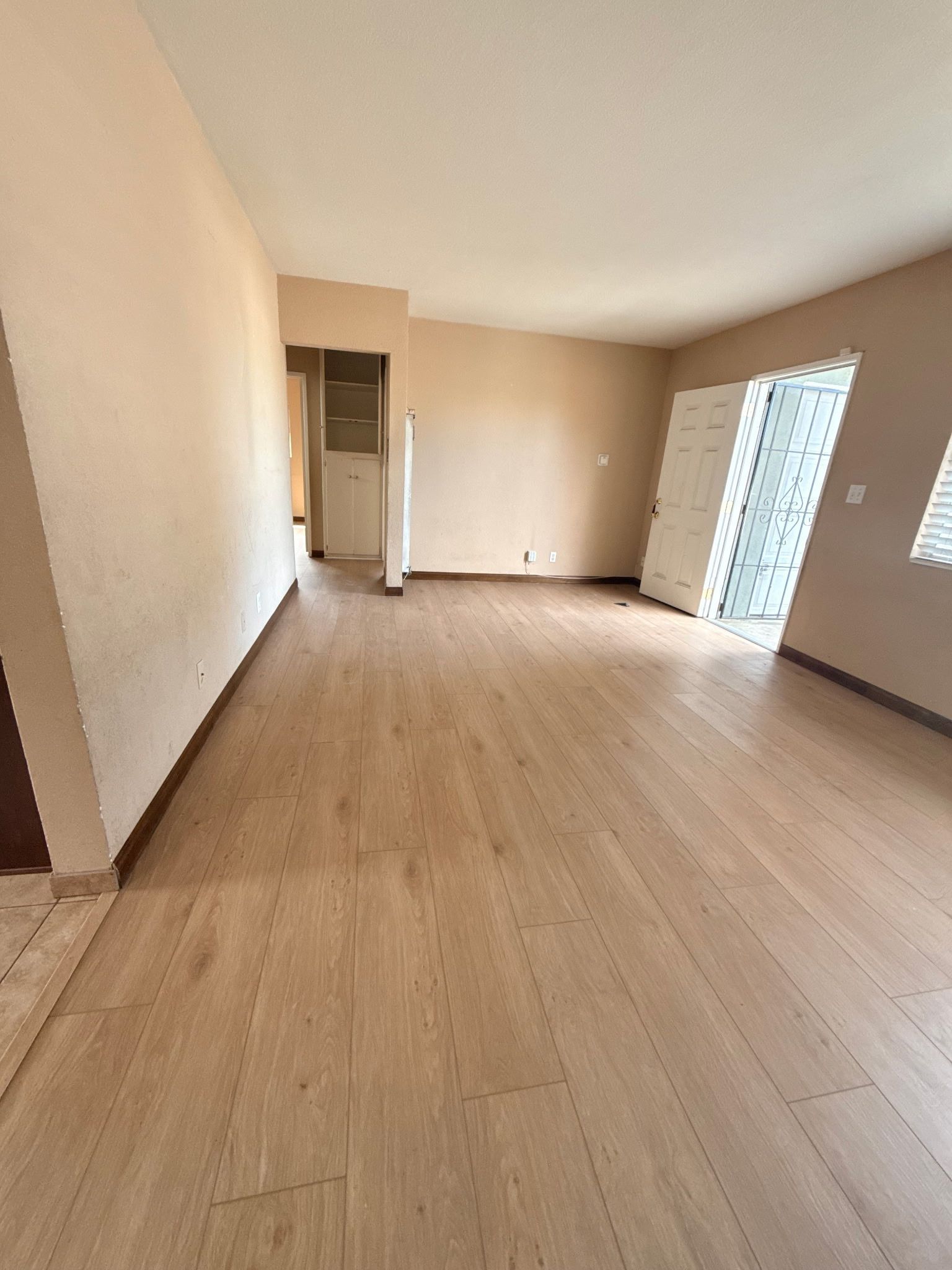 An empty room with light wood-look flooring, tan walls, a door, and a sliding glass door leading to a patio.