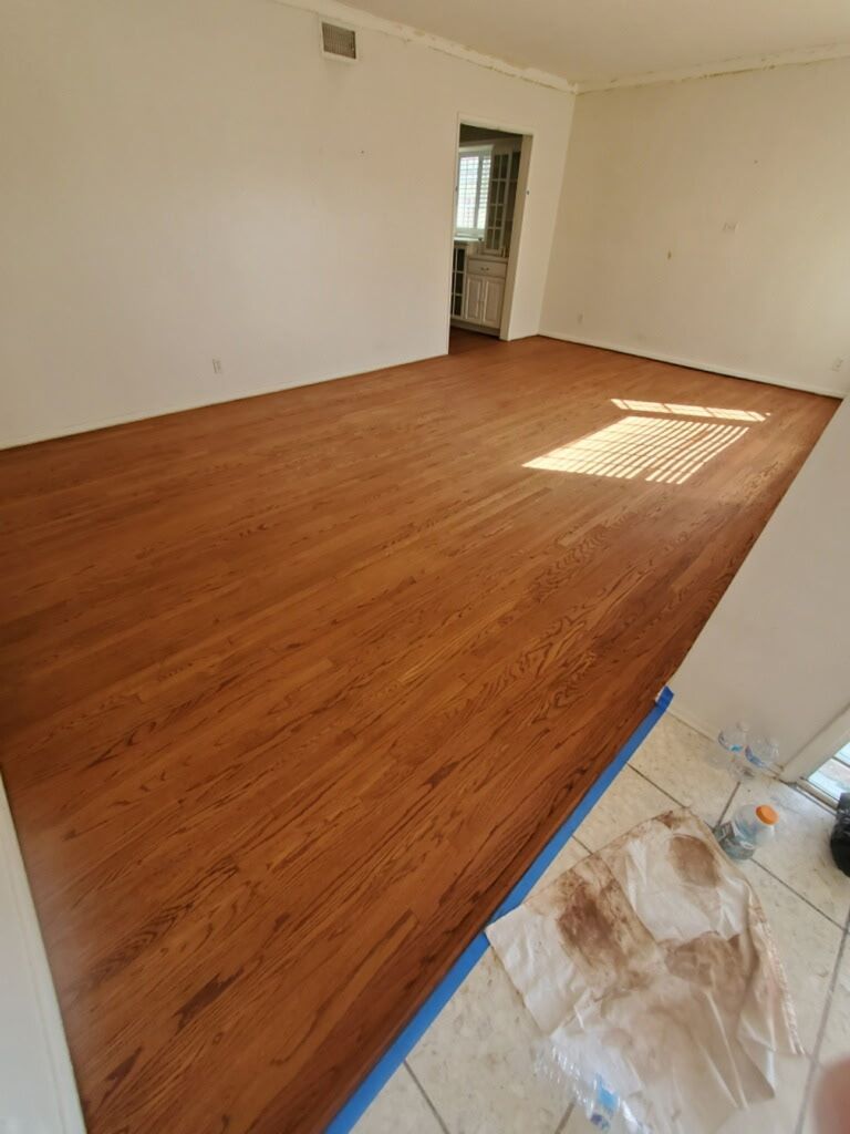 Newly refinished medium-brown hardwood floors in a room next to a tiled entryway with painting supplies.