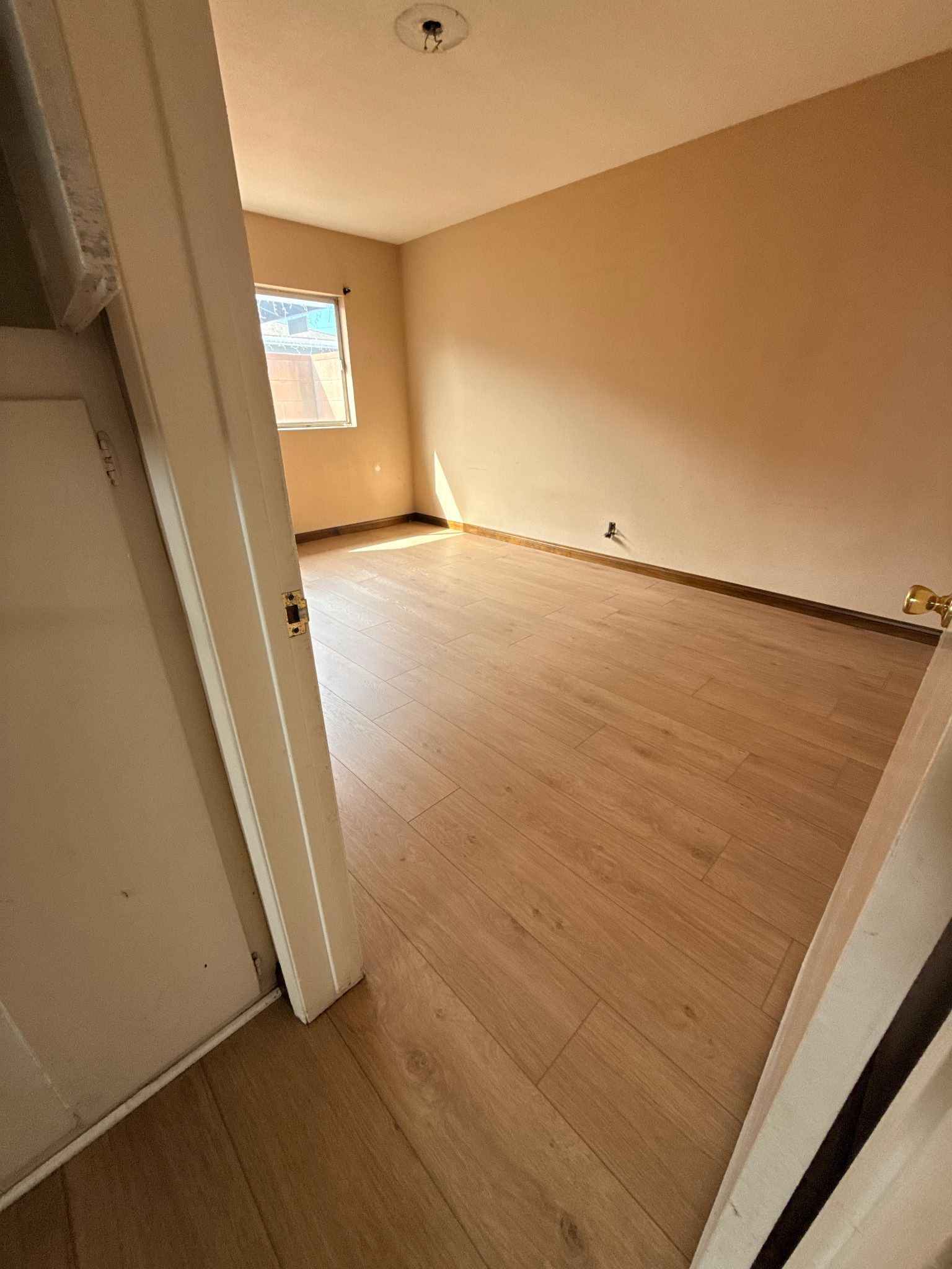 An empty room with light wood-look flooring, beige walls, a window on the far wall, and a ceiling light fixture.