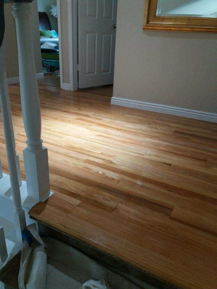 A polished hardwood floor in a brightly lit hallway, viewed from a staircase landing near a white wooden railing.