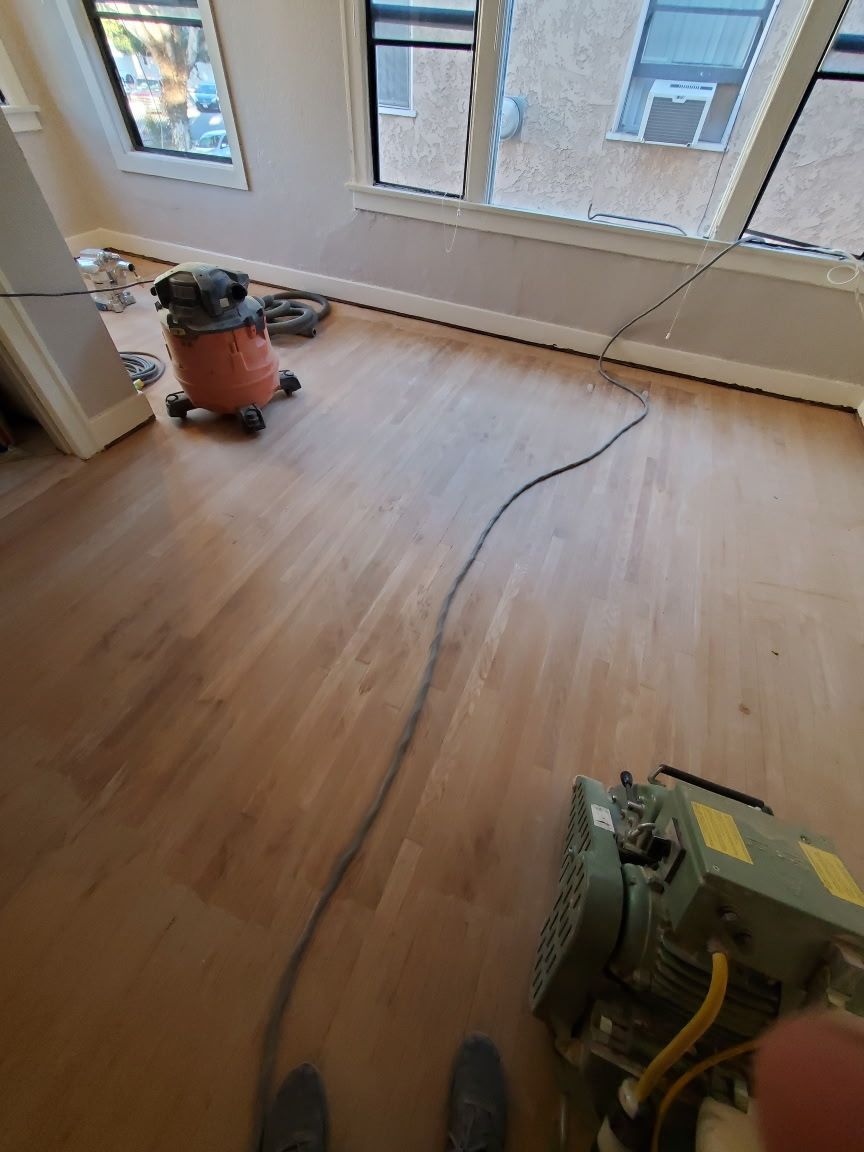 A hardwood floor being refinished with sanding equipment and a shop vacuum in a sunlit room.