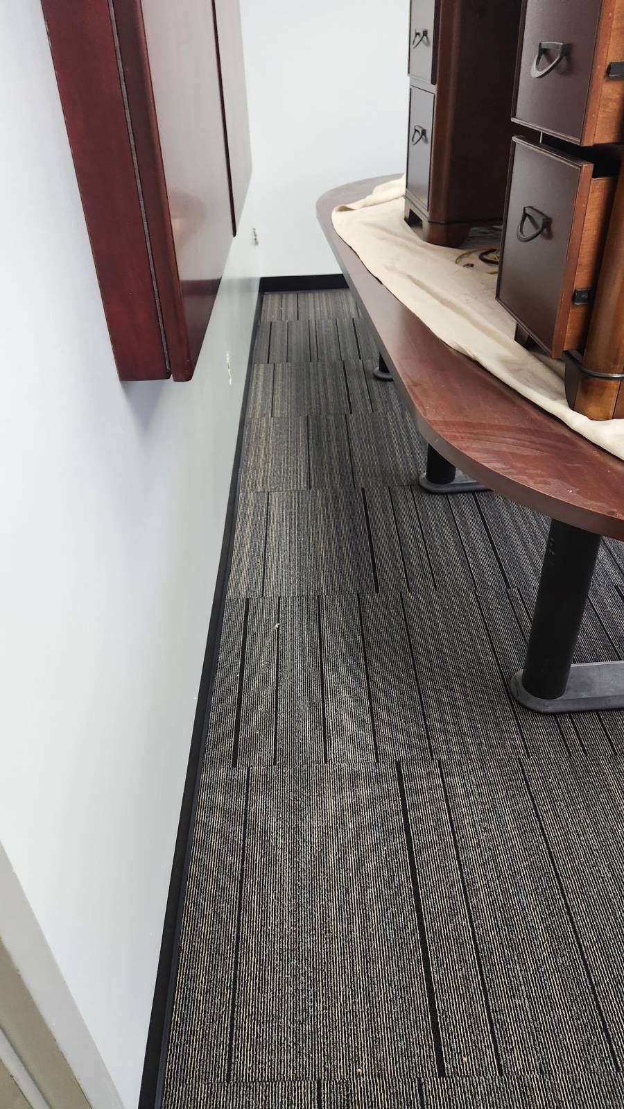 A high-angle view of a gray patterned carpet in a narrow office walkway beside a wooden desk and a dark wall.
