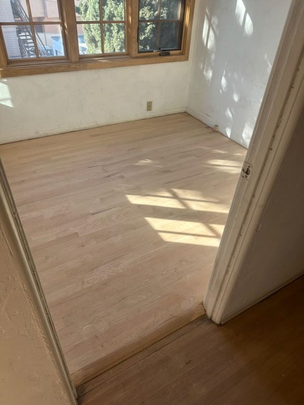 A room with newly sanded, unfinished light-wood flooring visible through a doorway, with bright window light casting shadows.