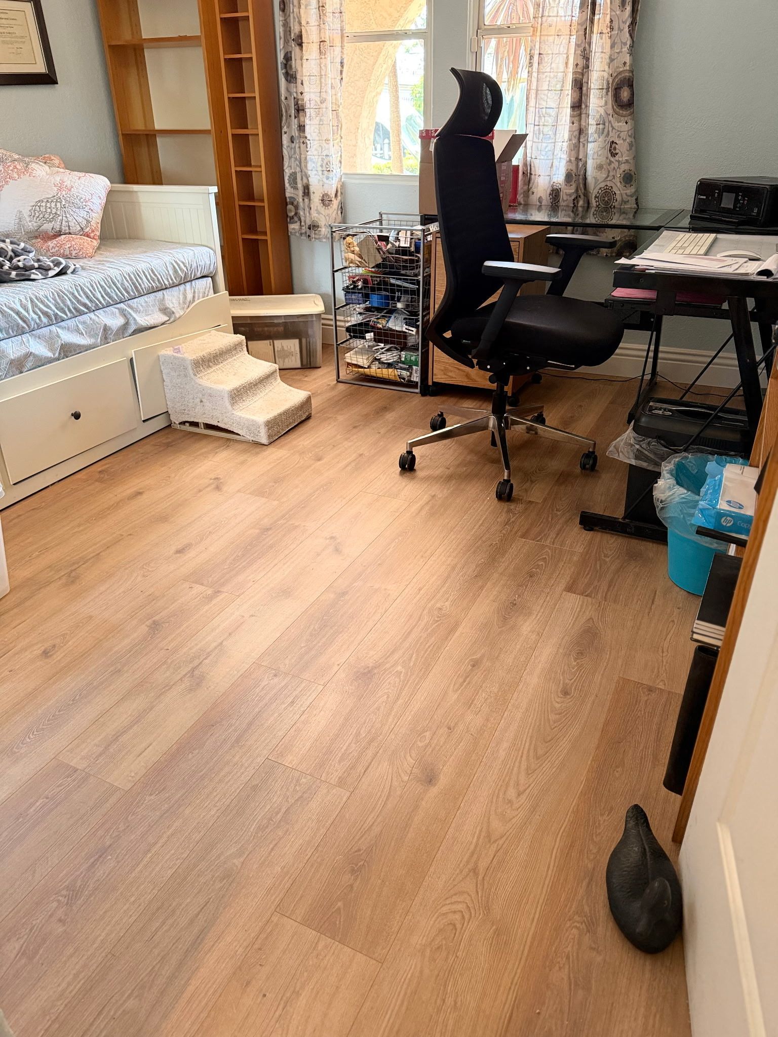A room featuring a white bed with steps, a wooden shelf, a desk with a black ergonomic chair, and light wood flooring.