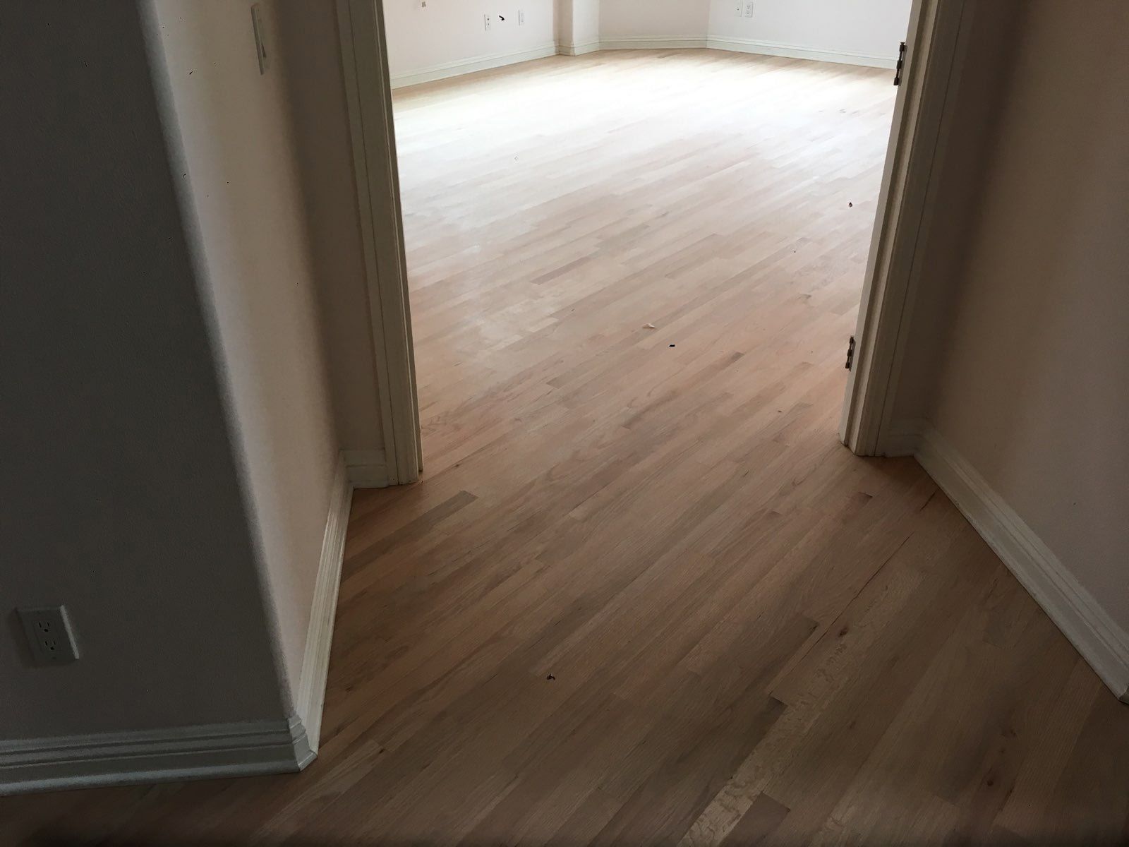 A wooden floor in a hallway leading into a brightly lit room through an open doorway with white trim and beige walls.