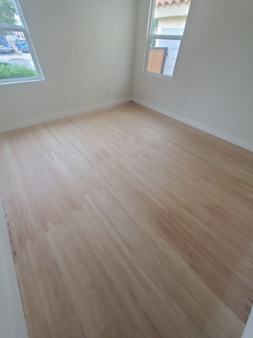 An empty room with light wood-tone flooring, white walls, and two windows allowing natural light.