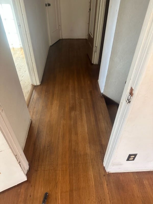 A narrow indoor hallway with light-reflecting wood flooring, flanked by white walls and several wooden doorways.