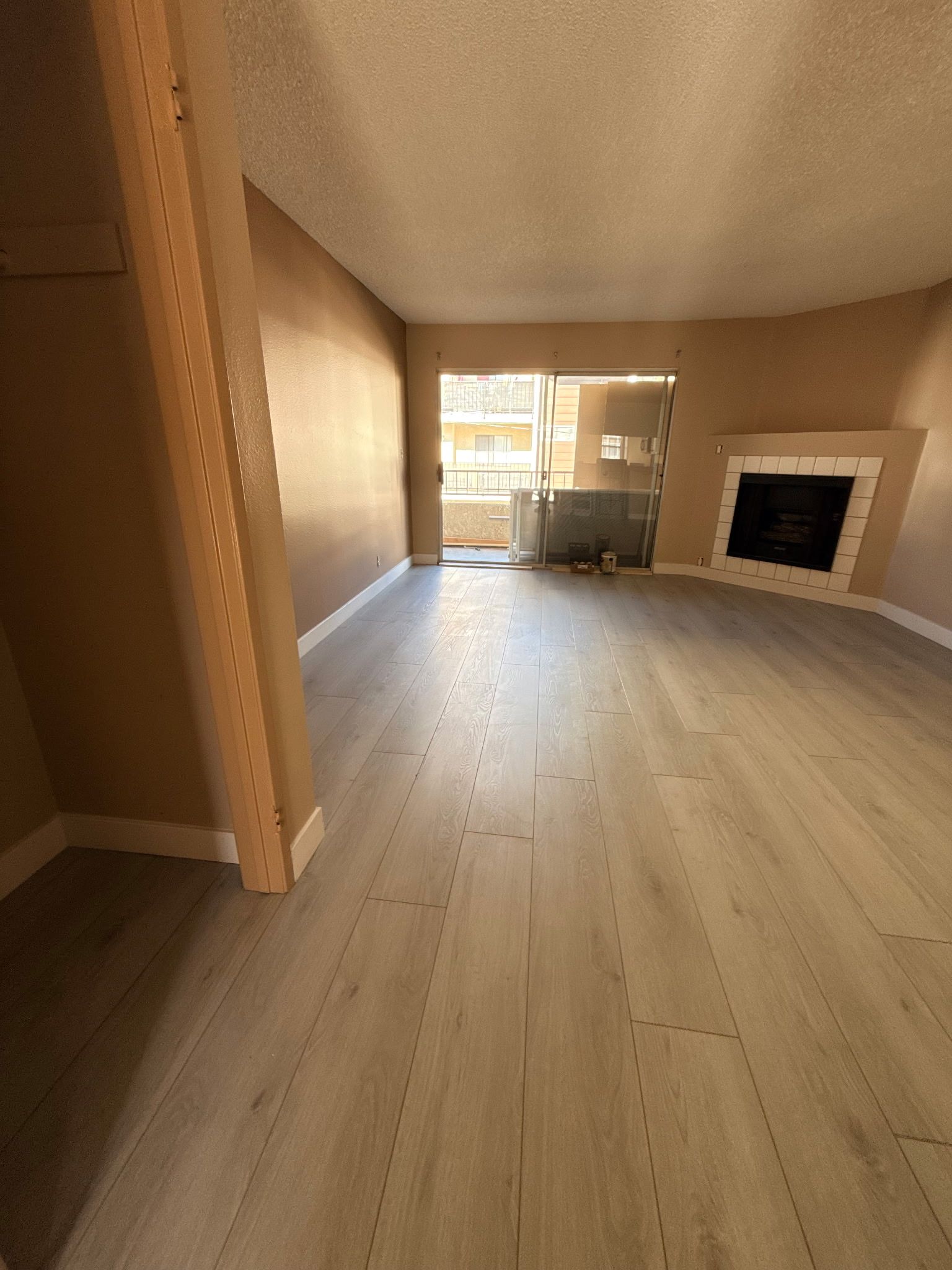 An empty room with light wood-look flooring, beige walls, a corner fireplace, and a glass door leading to a balcony.