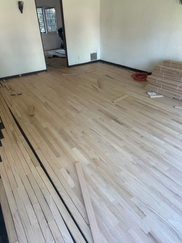 Unfinished light-colored wood flooring being installed in a room with a doorway and a stack of flooring boards nearby.