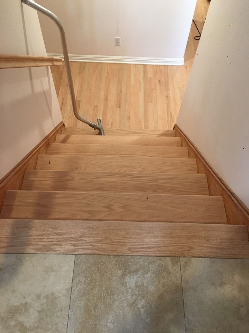 A high-angle view looking down a wooden staircase from a tiled landing to a hardwood floor, with a metal handrail.