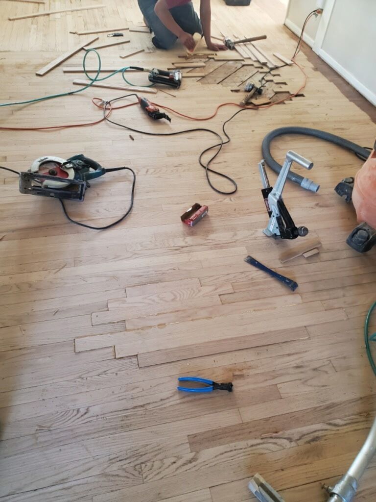 A person installs herringbone hardwood flooring in a room filled with various woodworking power tools and equipment.
