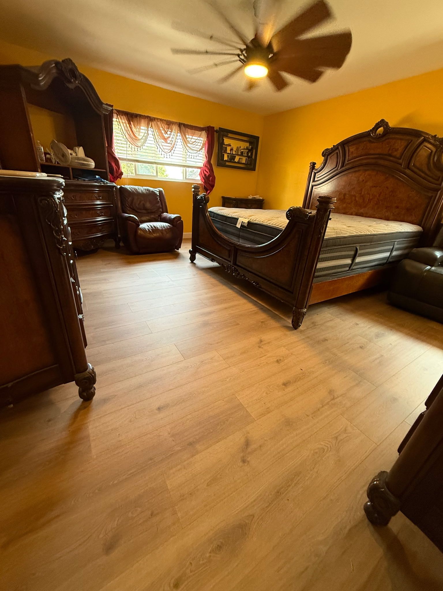 A bedroom with mustard yellow walls, hardwood floors, a large ornate wood bed frame, a matching dresser, and an armchair.