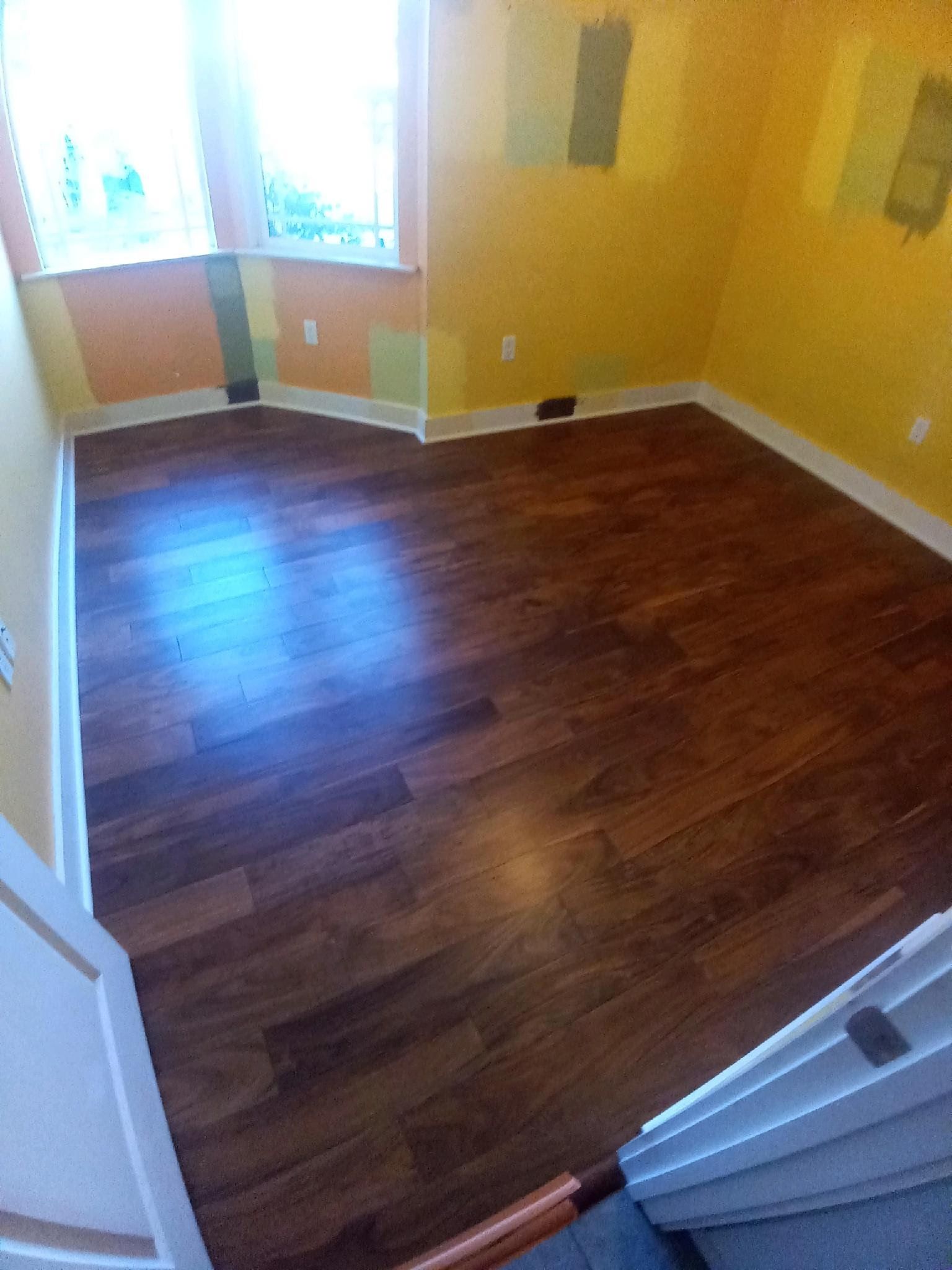 A bright room with new dark wood flooring and yellow walls showing paint swatches around a bay window.