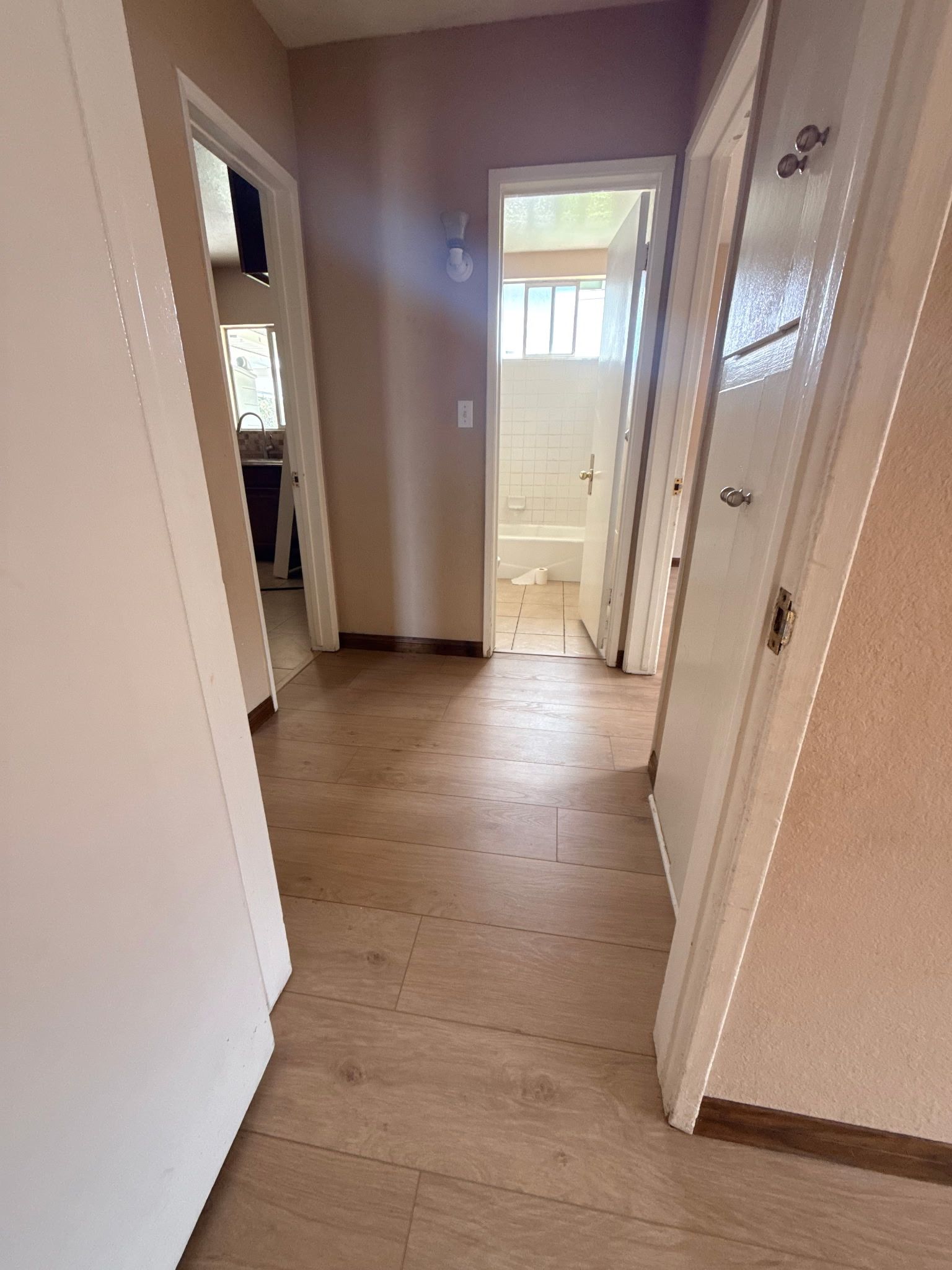 A hallway with light wood-look flooring leading to a bathroom doorway, with additional doors on the sides.