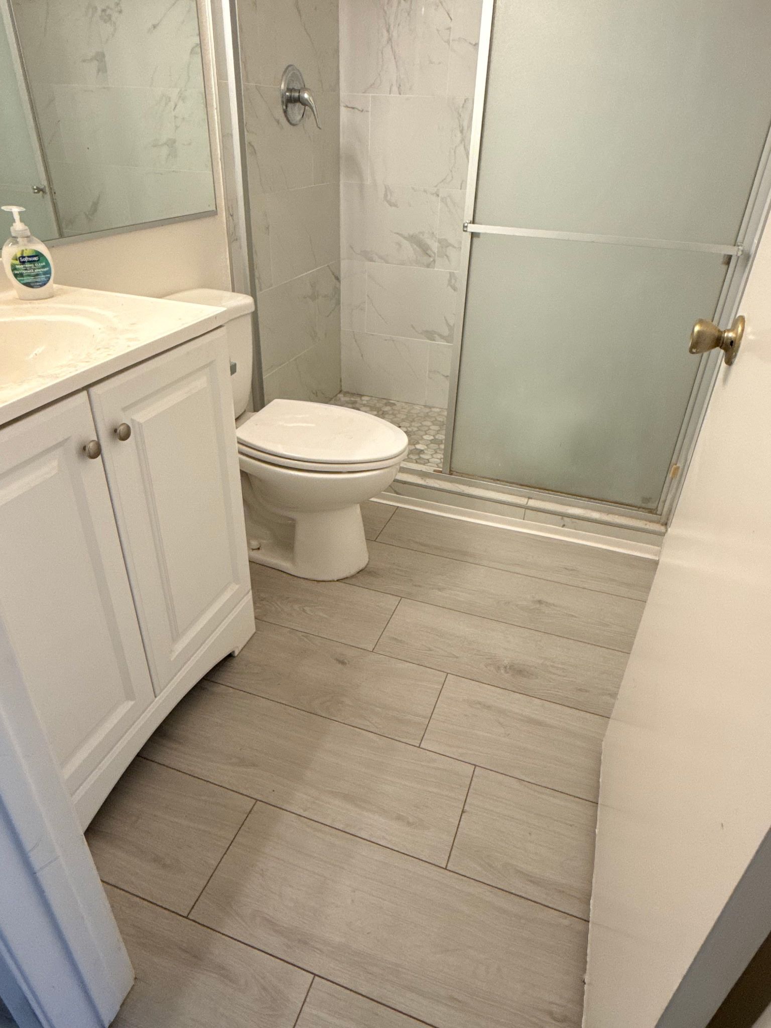 A small bathroom with a white vanity, a white toilet, and a glass-enclosed shower on grey tiled flooring.