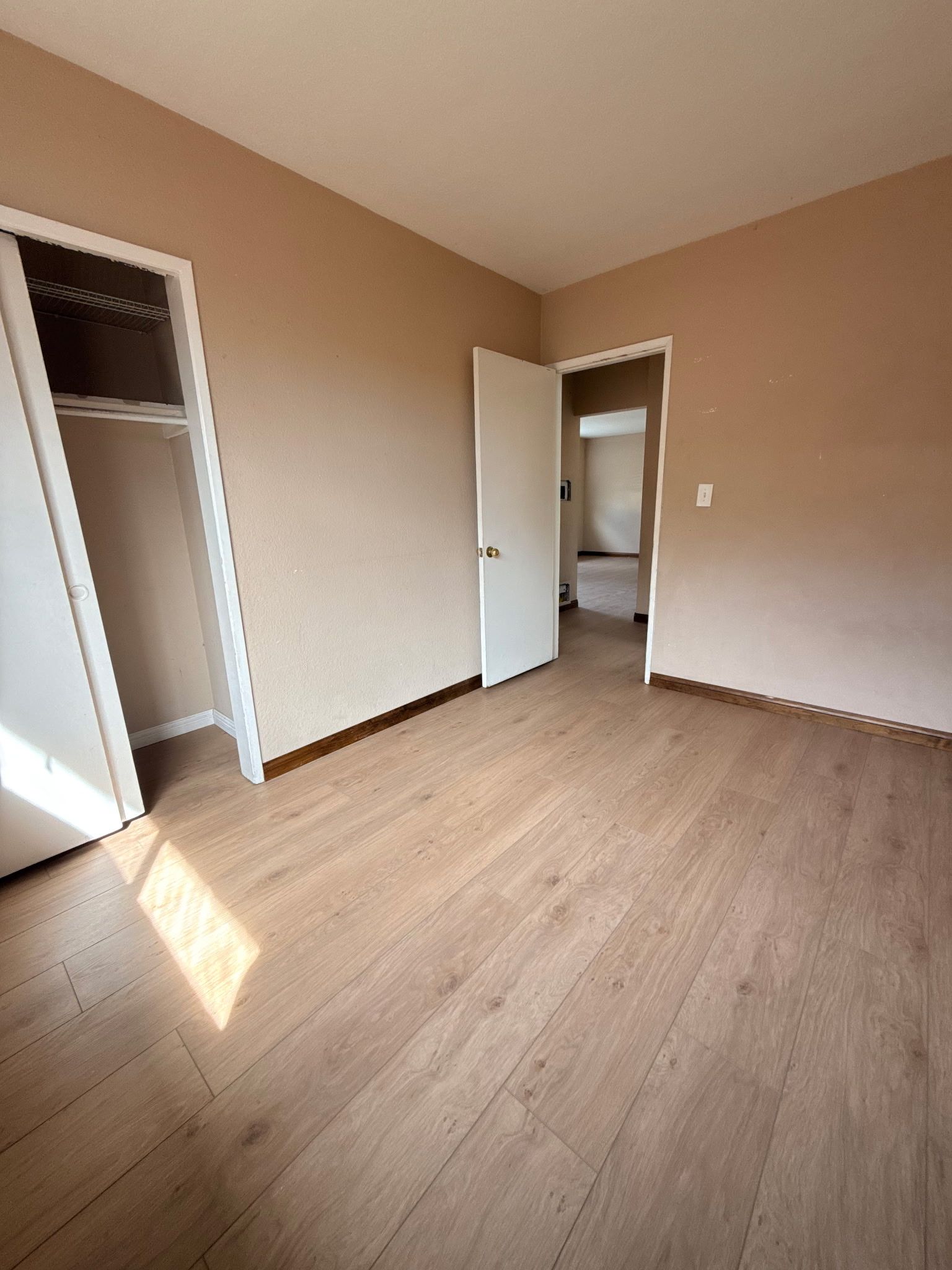 An empty room with light wood-look flooring, tan walls, a closet, and an open doorway leading to another room.