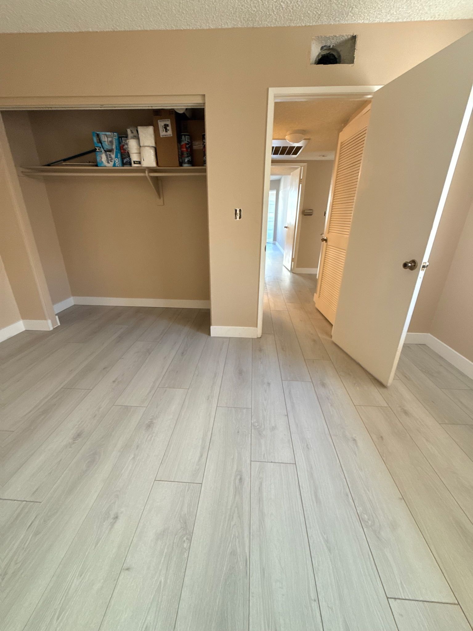 A bedroom interior with light wood floors, a closet containing shelving and items, and an open door leading to a hallway.