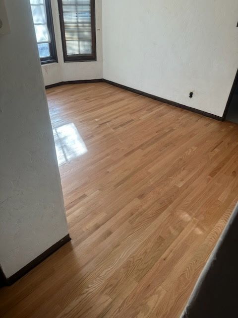 An empty room featuring light wood floors, white walls with dark baseboards, and windows on the far wall.