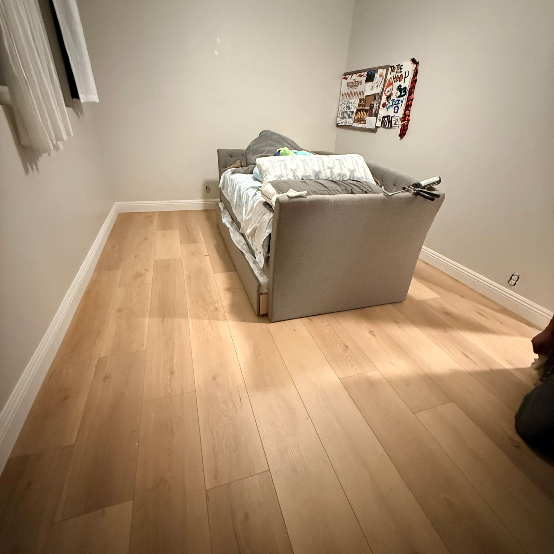 A gray daybed sits in the center of an empty bedroom with light wood flooring, white walls, and a wall collage.