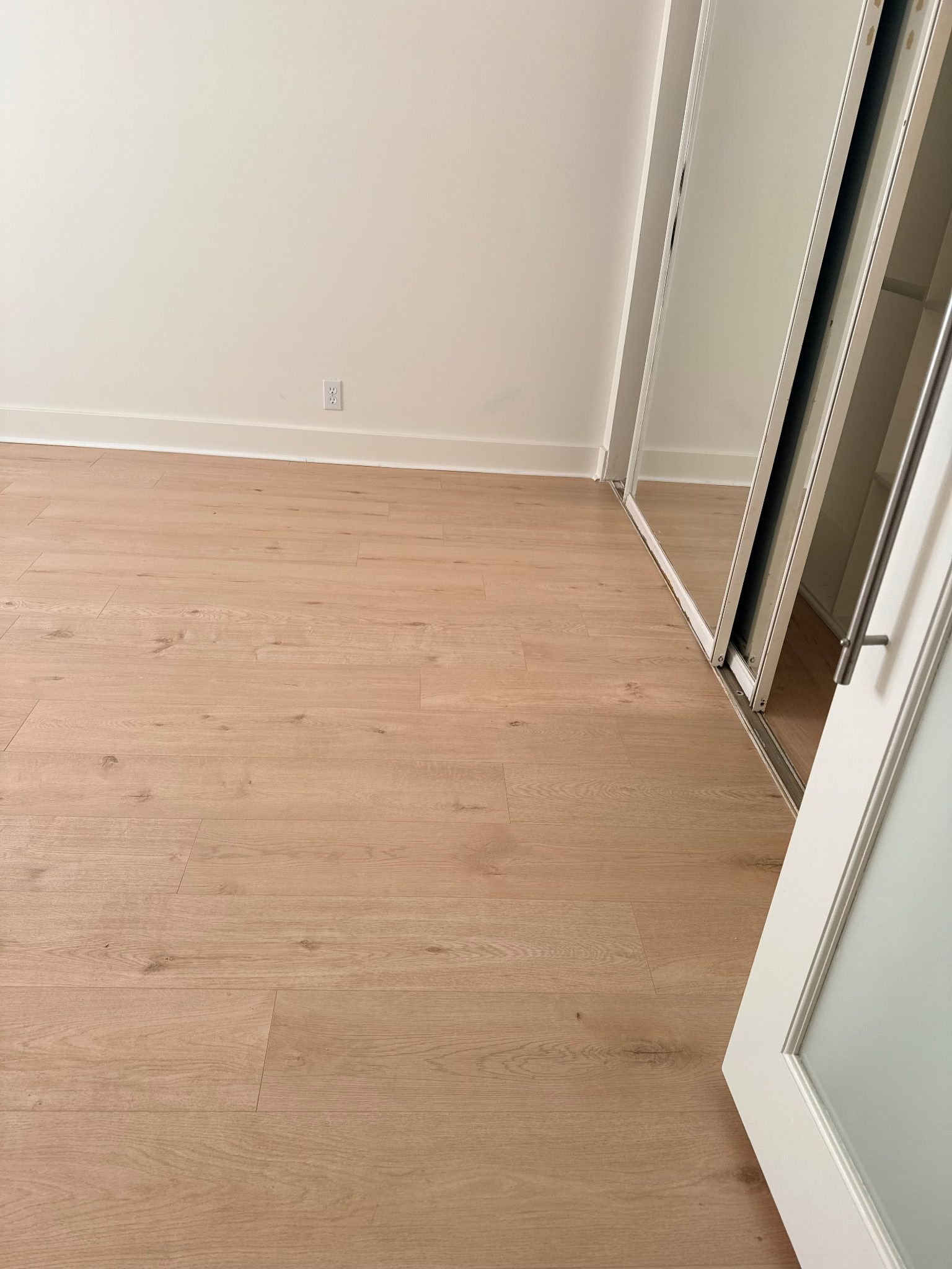 A view of a room with light-colored hardwood flooring, white walls, and a large mirrored closet door on the right.