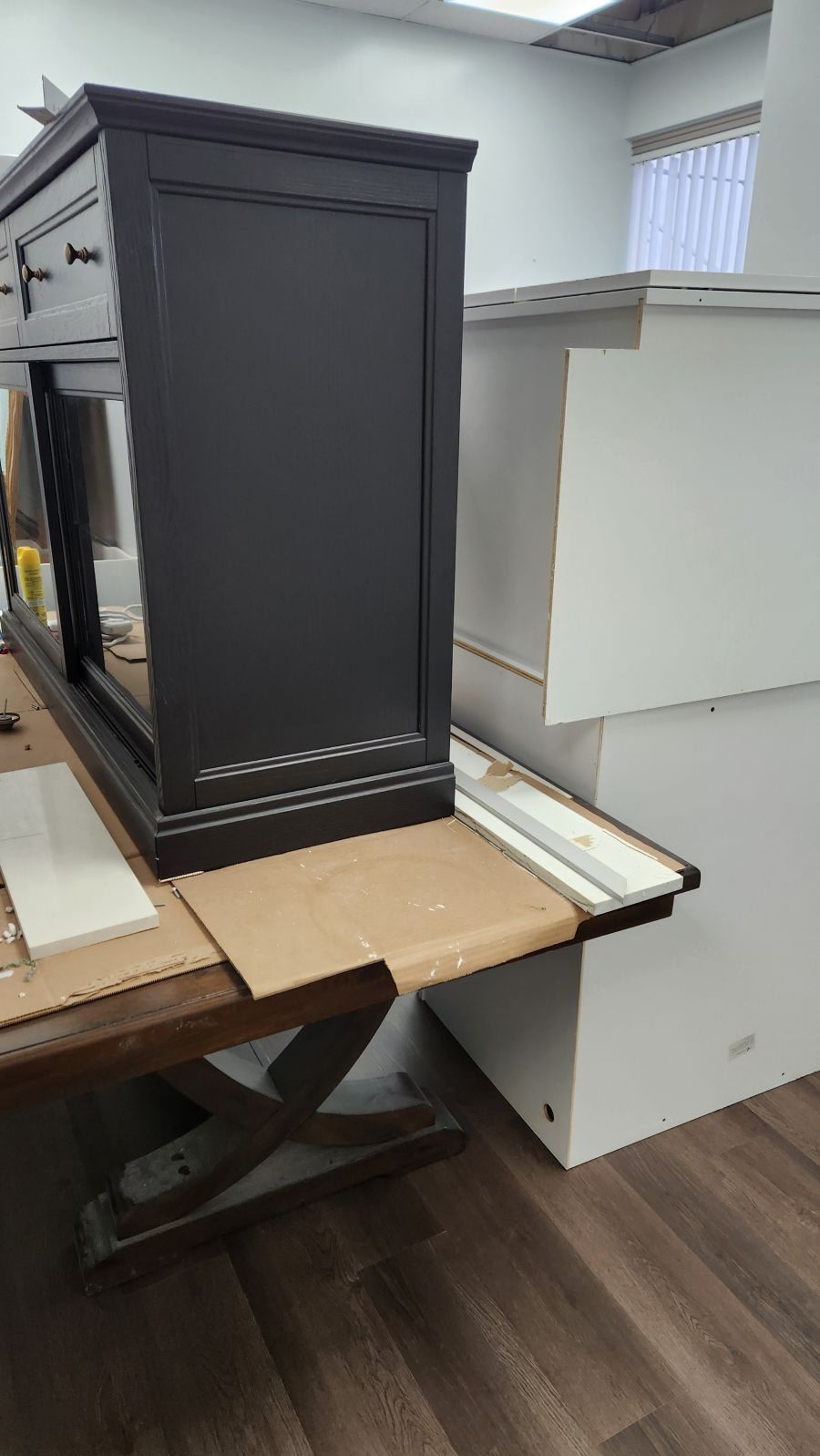 A dark gray dresser sits on a workbench next to a white cabinet piece in a workshop with light wood-look flooring.