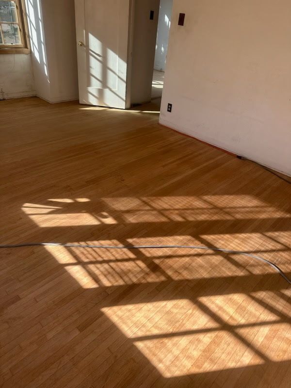 A high-angle view of a sunlit room with hardwood floors, showing window pane shadows cast across the wooden planks.