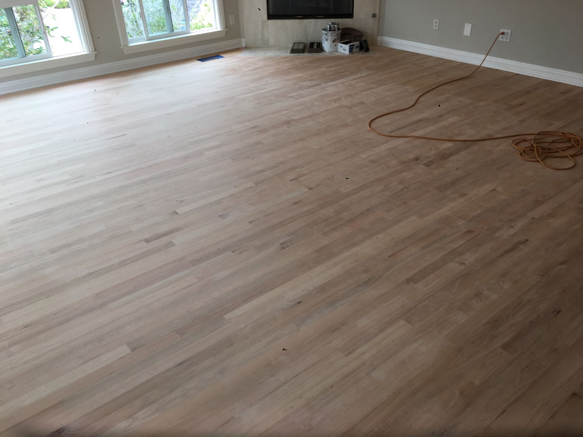 Newly sanded, unfinished oak hardwood flooring in a room with a corner fireplace and windows.