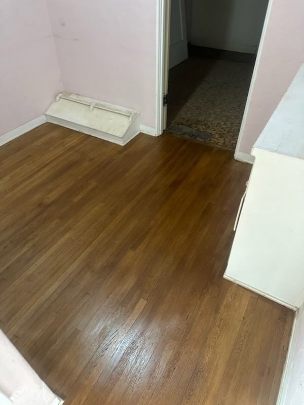 A room with hardwood flooring, a white baseboard heater against a light pink wall, and a doorway leading to a carpeted hall.