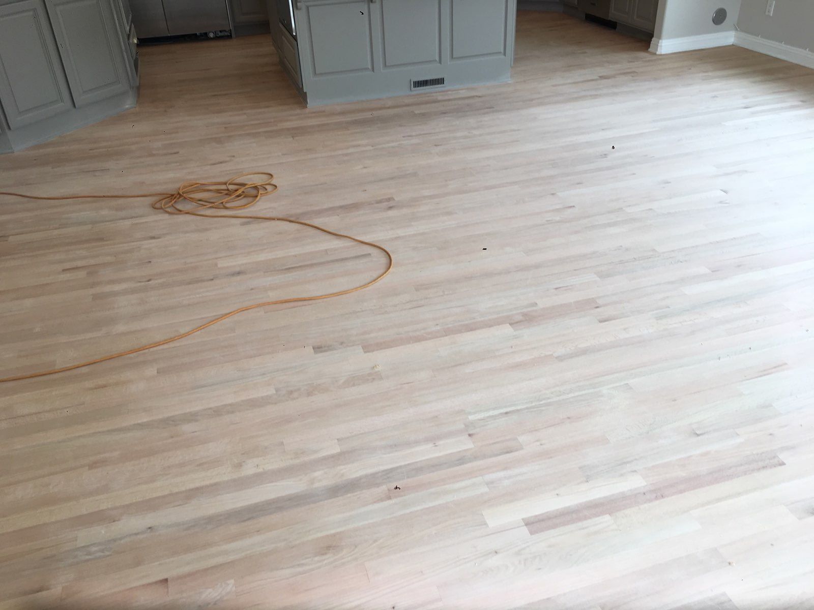 A newly sanded, light-colored hardwood floor in a kitchen with gray cabinets and a coiled extension cord on the floor.