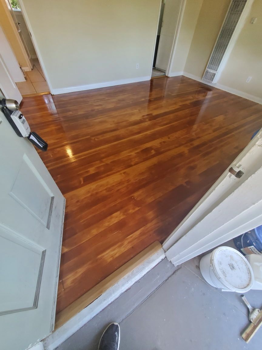 A view through a doorway into a room with freshly refinished, glossy wood flooring and neutral walls.