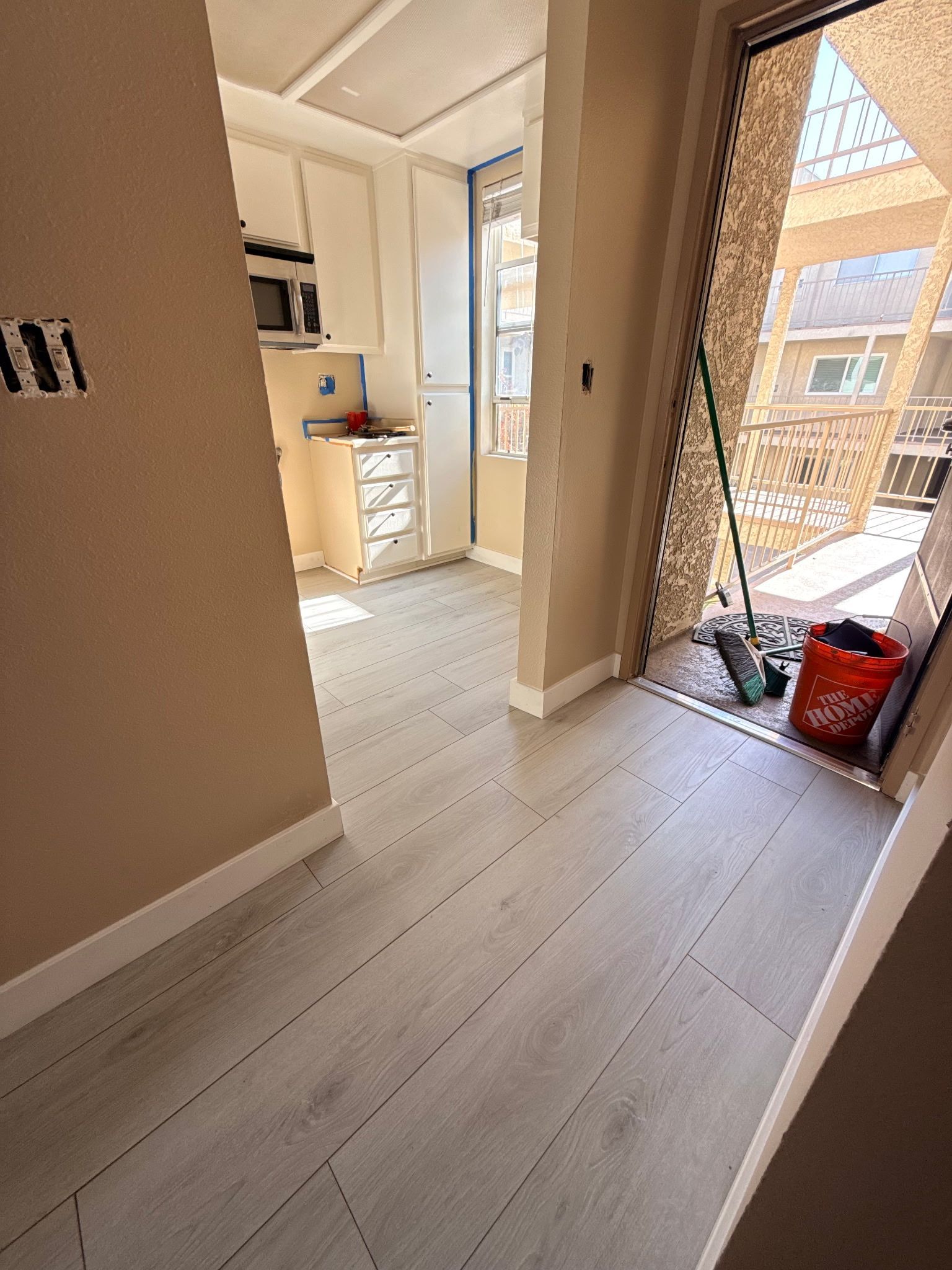 A view from a doorway looking into a kitchen with light wood-style flooring, white cabinets, and a red bucket near the exit.