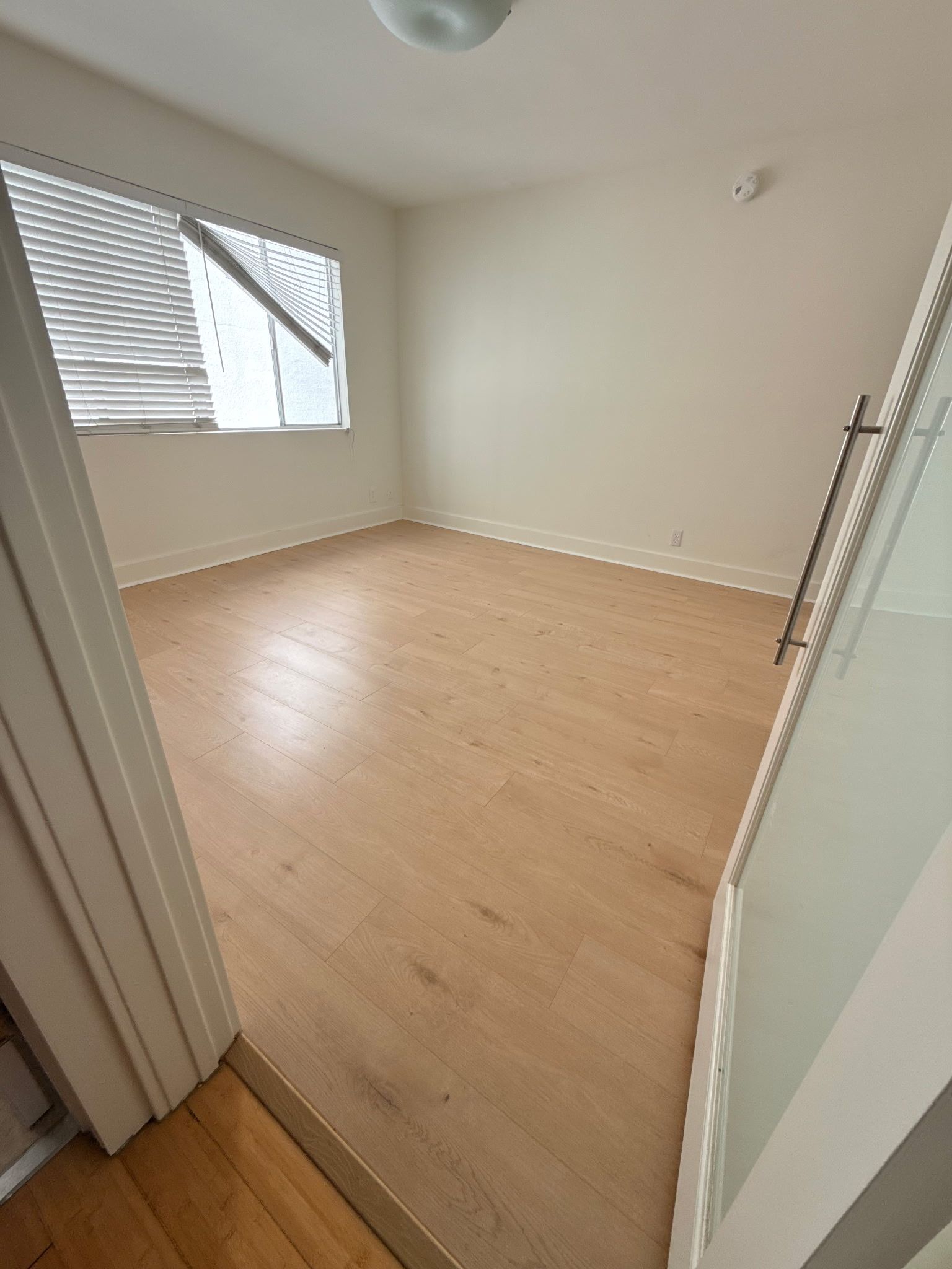 An empty room with light wood-look flooring, white walls, and a window with white blinds.