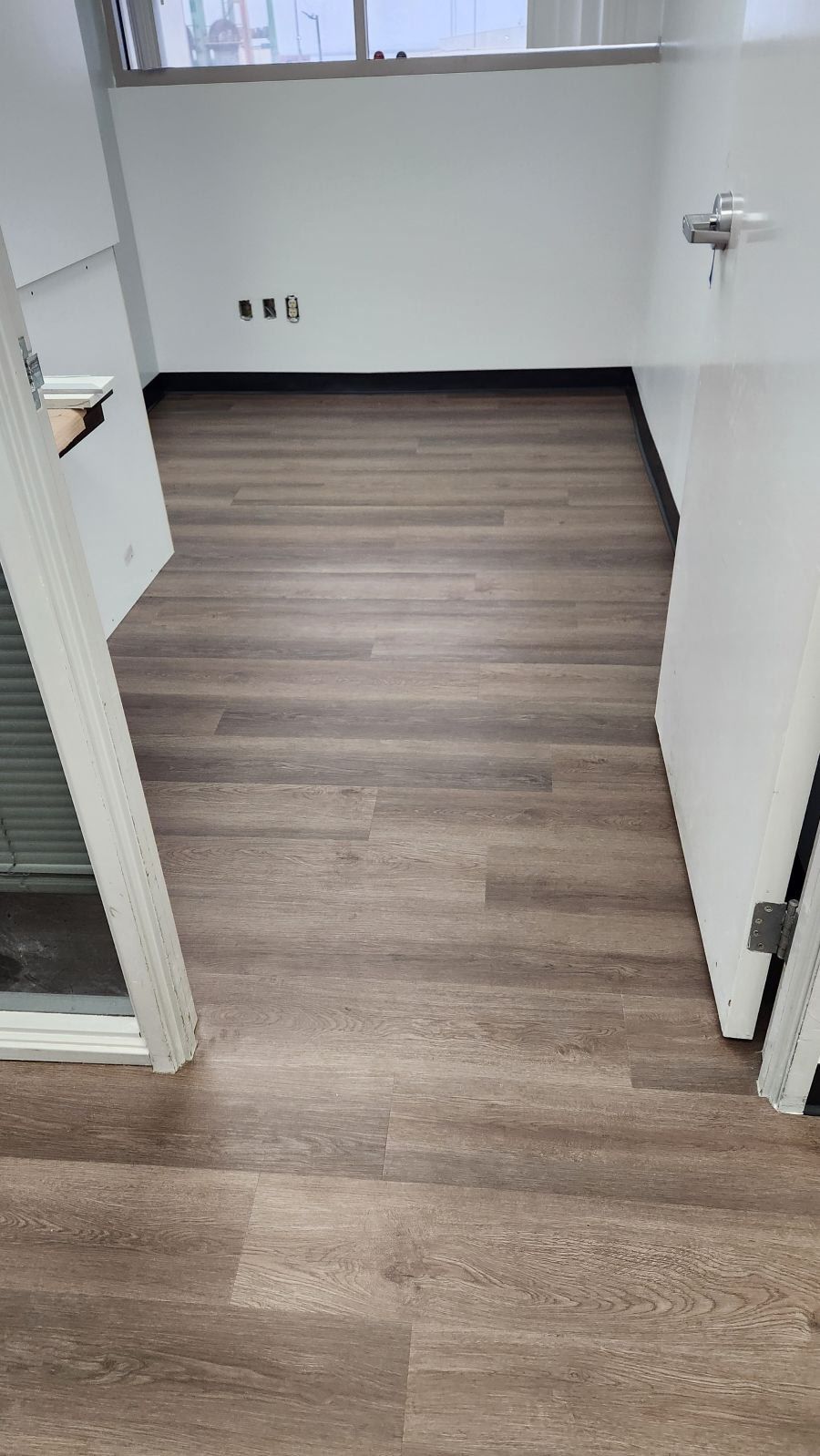 An empty office room with light grey walls, a window, and wood-patterned vinyl plank flooring.