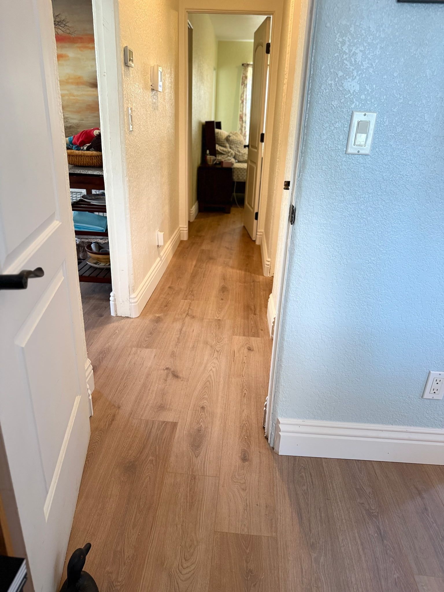 A hallway featuring wood-look flooring, white trim, and textured blue and beige walls, leading to an open bedroom doorway.