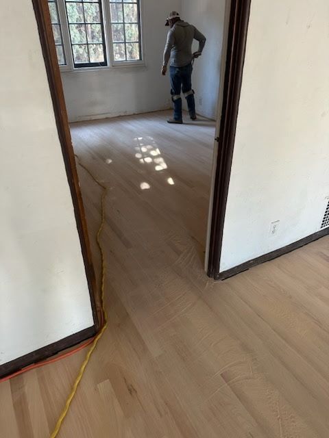 A worker in a room with light-colored, newly sanded hardwood flooring, seen through an open doorway.