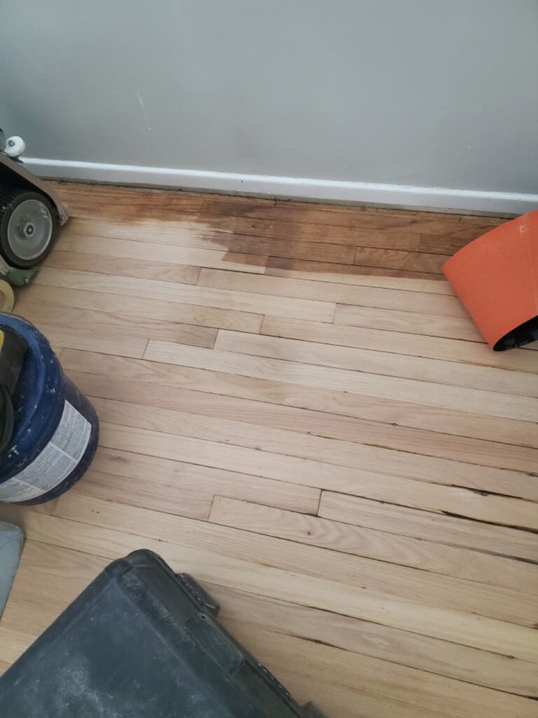 A partial view of a hardwood floor being sanded, showing both raw light wood and a strip of darker, unfinished wood.