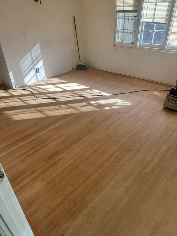 An interior view of a room with light-colored, newly sanded hardwood flooring and plain white walls under bright lighting.