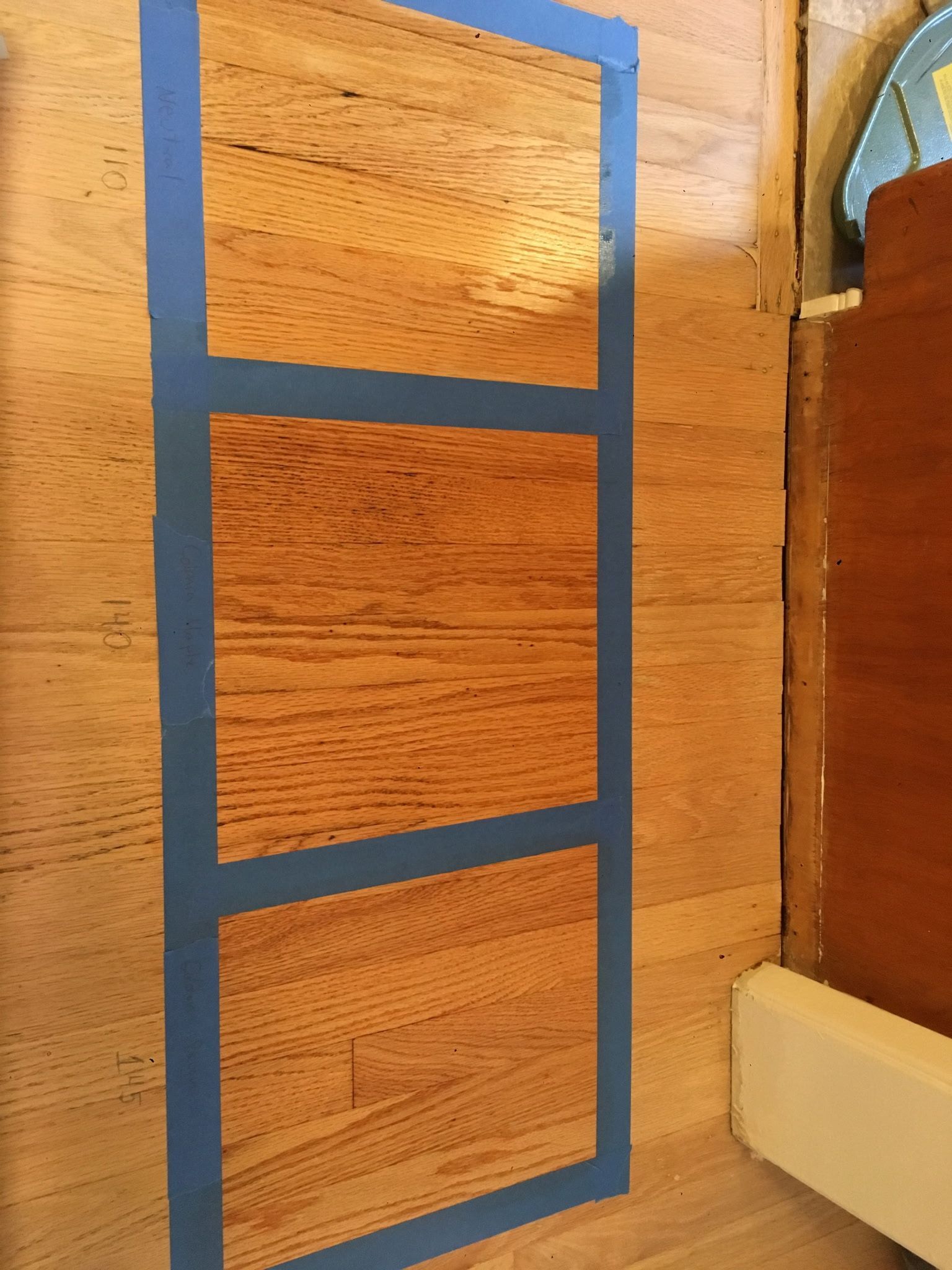 Three test patches of wood stain on hardwood flooring, outlined with blue painter’s tape to compare different shades.
