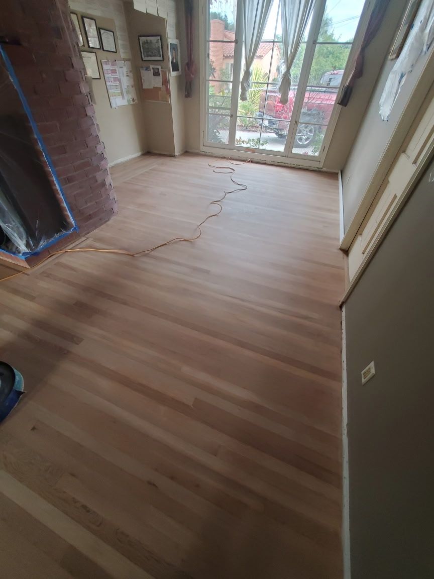 A room with light-colored, newly refinished hardwood floors, a brick fireplace, and large windows looking outdoors.