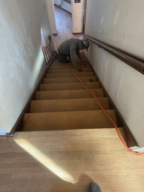 A person in a gray shirt works on wood-paneled stairs, with an orange power cord extending down the staircase.