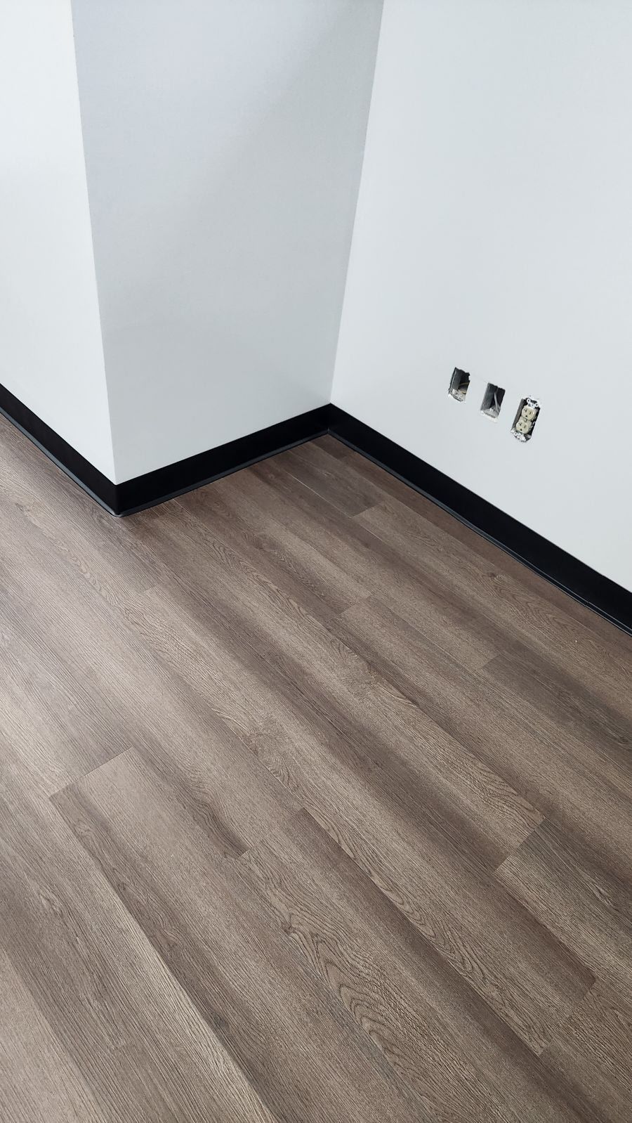 A corner of a room with white walls, black baseboards, wood-look flooring, and three wall outlets.