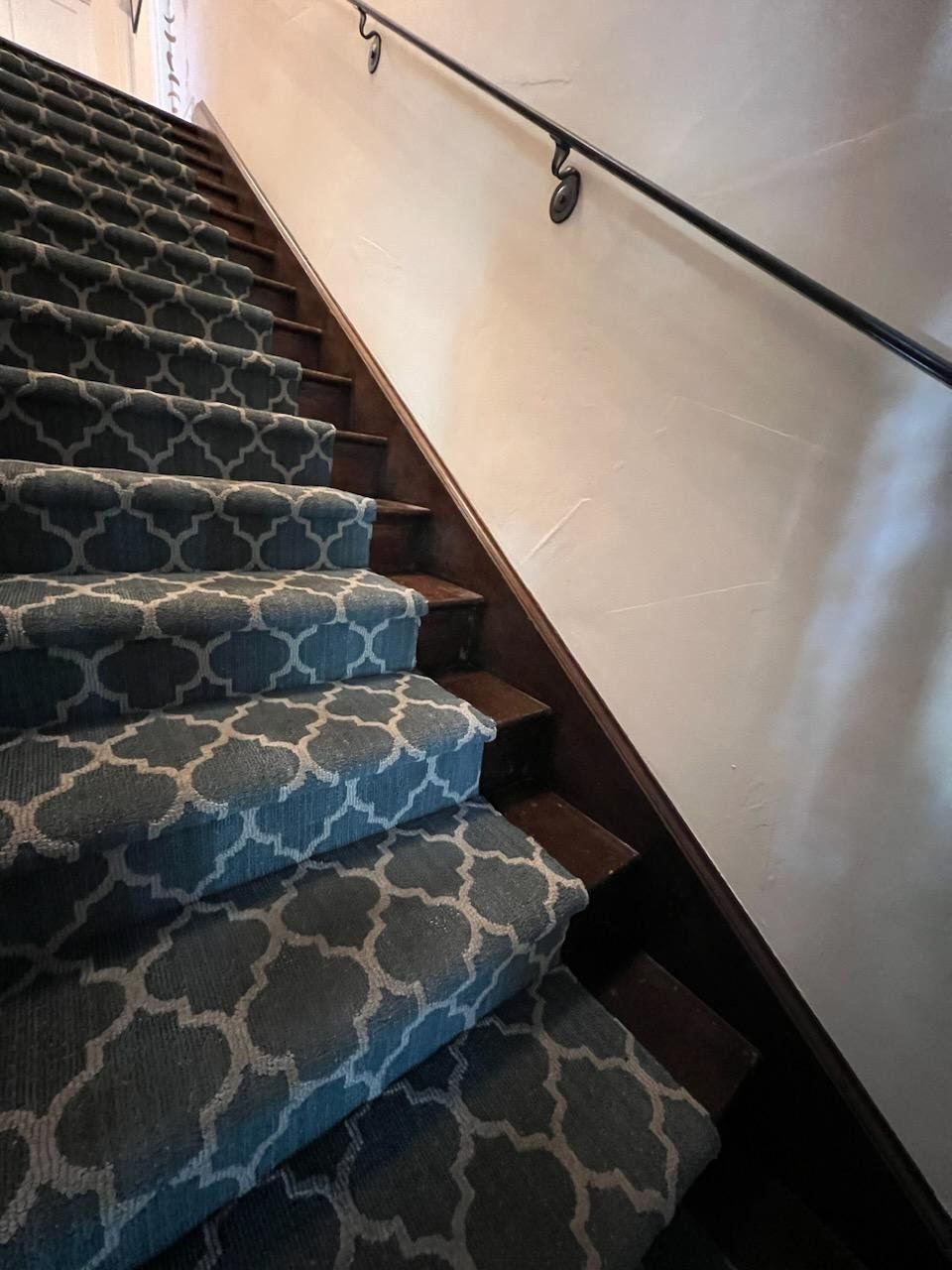 A staircase with dark wood treads and a blue-and-white lattice-patterned carpet runner, next to a white wall with a railing.