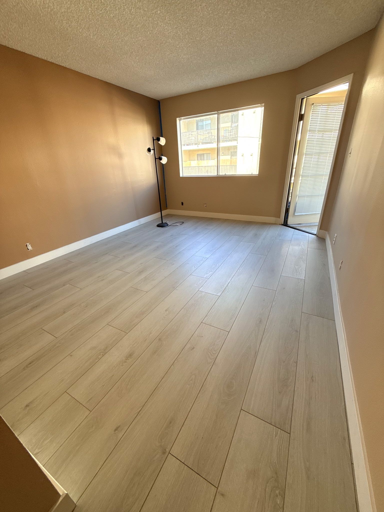 Empty living room with light wood-look flooring, beige walls, a floor lamp, and an open doorway leading outdoors.